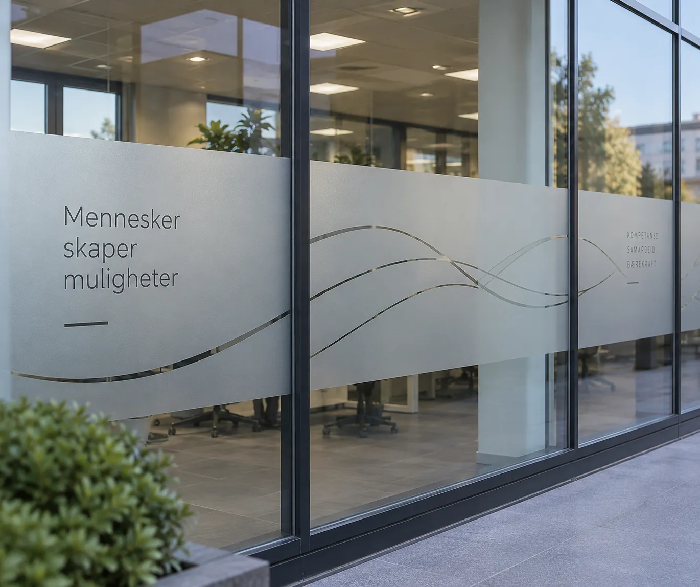 Frostet foliedekor med bølgende linjer og teksten «Mennesker skaper muligheter» på en glassfasade
